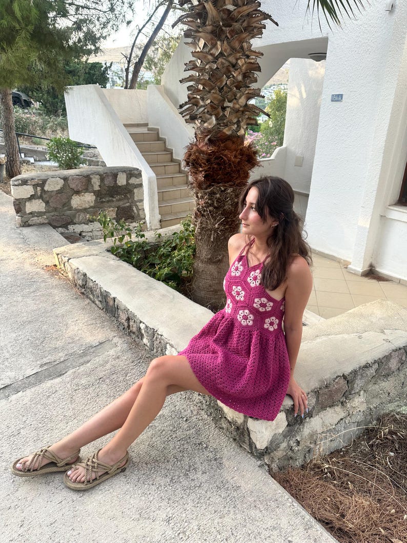 May include: A magenta crochet dress with white floral detailing. The dress has a halter neck and a flared skirt. The image also shows beige sandals with woven straps. The background includes a stone wall, steps, and a palm tree.