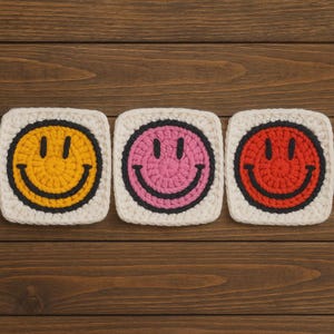 Könnte beinhalten: Drei quadratische Häkeluntersetzer mit Smiley-Gesichtern. Jeder Untersetzer hat ein andersfarbiges Smiley-Gesicht: gelb, rosa und rot, schwarz umrandet, auf weißem Hintergrund. Die Untersetzer sind auf einer Holzoberfläche angeordnet.
