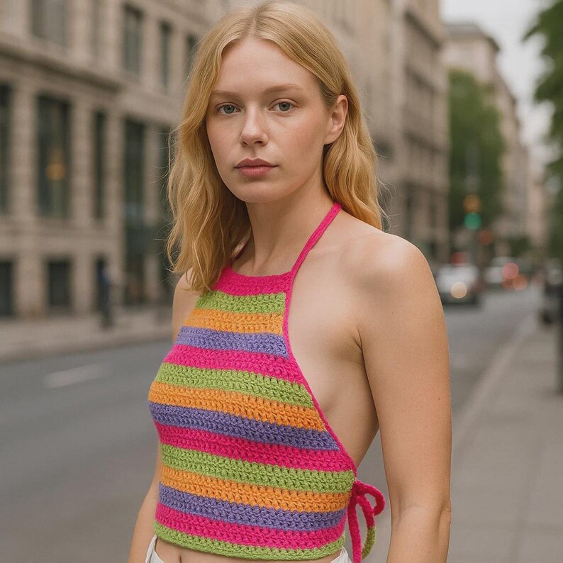 K&ouml;nnte beinhalten: Ein bunt gestreiftes Halterneck-Top im H&auml;kelstil. Das Top hat horizontale Streifen in Pink, Gr&uuml;n, Orange und Lila. Die Haltertr&auml;ger und Seitenschlaufen sind einfarbig pink. Das Top wird im Freien getragen.