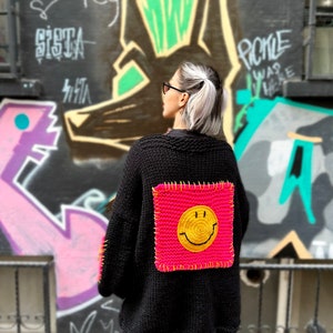 Puede incluir: C&aacute;rdigan de punto negro con un gran parche cuadrado rosa ne&oacute;n con una carita sonriente amarilla en la espalda. El c&aacute;rdigan tiene mangas largas. El fondo presenta arte de graffiti.