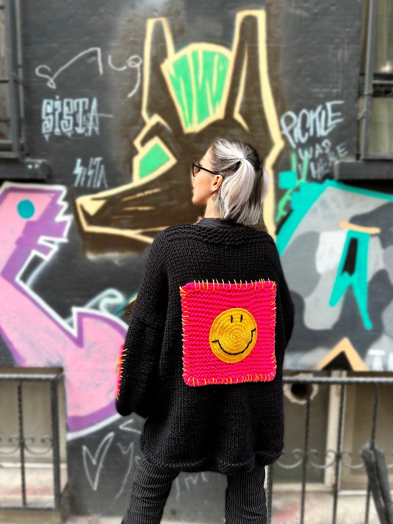 Puede incluir: C&aacute;rdigan de punto negro con un gran parche cuadrado rosa ne&oacute;n con una carita sonriente amarilla en la espalda. El c&aacute;rdigan tiene mangas largas. El fondo presenta arte de graffiti.