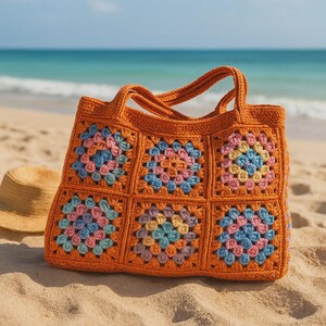 Puede incluir: Un bolso tote de crochet naranja con cuadrados florales coloridos. El bolso tiene dos asas y est&aacute; sobre una playa de arena con un sombrero de paja cerca. Los cuadrados presentan detalles en rosa, azul y amarillo.