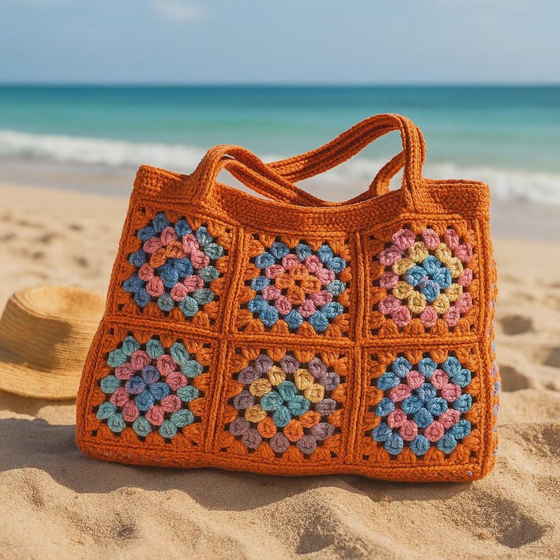 Puede incluir: Un bolso tote de crochet naranja con cuadrados florales coloridos. El bolso tiene dos asas y est&aacute; sobre una playa de arena con un sombrero de paja cerca. Los cuadrados presentan detalles en rosa, azul y amarillo.