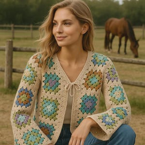 Könnte beinhalten: Beige Häkel-Cardigan mit V-Ausschnitt und Bindverschluss. Der Cardigan hat bunte quadratische Muster in Blau-, Grün-, Lila- und Brauntönen. Die Ärmel sind lang und ausgestellt. Der Cardigan wird mit einer blauen Jeans getragen.