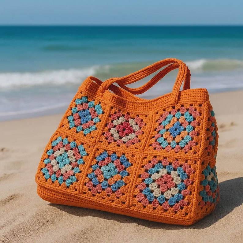 Puede incluir: Un bolso tote de crochet naranja con un dise&ntilde;o de cuadrados de la abuela. El bolso presenta cuadrados coloridos en tonos rosa, azul y crema. Tiene dos asas y est&aacute; sobre una playa de arena, con el oc&eacute;ano al fondo.