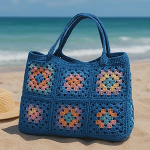 Puede incluir: Un bolso tote de crochet azul con cuadrados florales coloridos. El bolso tiene dos asas y est&aacute; sobre una playa de arena con el oc&eacute;ano de fondo. Los cuadrados florales son en tonos rosa, naranja y amarillo.