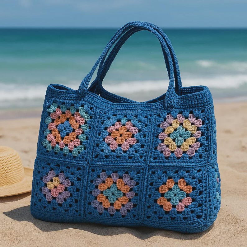 Puede incluir: Un bolso tote de crochet azul con cuadrados florales coloridos. El bolso tiene dos asas y est&aacute; sobre una playa de arena con el oc&eacute;ano de fondo. Los cuadrados florales son en tonos rosa, naranja y amarillo.