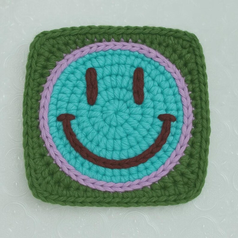 May include: A crocheted square with a smiley face design. The face is turquoise with brown eyes and a smile, encircled by a lavender ring and a green border. This handmade item could be a coaster or decorative piece.