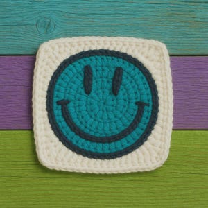 May include: A crocheted smiley face design in a square shape. The face is teal with a dark teal outline, featuring a smiling mouth and two eyes. The face is set on a white square with a crocheted border. The background is a wooden surface with teal, purple, and green planks.