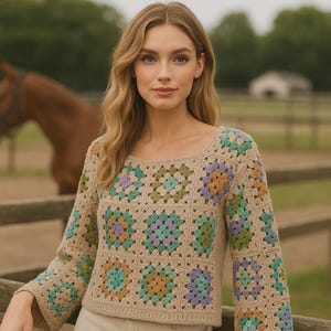 Könnte beinhalten: Ein beigefarbener Häkelpullover mit quadratischem Ausschnitt und langen Ärmeln. Der Pullover hat ein farbenfrohes Patchwork-Design mit Quadraten in Grün-, Blau-, Lila- und Orangetönen. Der Hintergrund zeigt ein Pferd und einen Holzzaun.