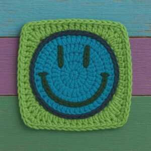 May include: A square, crocheted coaster with a smiley face design. The face is blue with a dark blue outline and a green smile. The coaster has a green border and is set against a colourful wooden background.