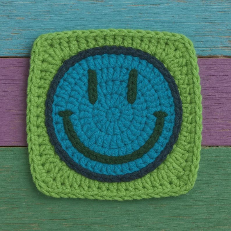 May include: A square, crocheted coaster with a smiley face design. The face is blue with a dark blue outline and a green smile. The coaster has a green border and is set against a colourful wooden background.
