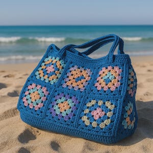 Puede incluir: Un bolso tote de crochet azul con un dise&ntilde;o de cuadrados de la abuela. El bolso presenta una base y asas azules lisas, con cuadrados coloridos en tonos pastel de rosa, naranja y morado. El bolso est&aacute; sobre una playa de arena con el oc&eacute;ano al fondo.