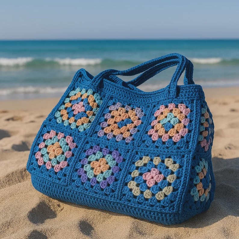 Puede incluir: Un bolso tote de crochet azul con un dise&ntilde;o de cuadrados de la abuela. El bolso presenta una base y asas azules lisas, con cuadrados coloridos en tonos pastel de rosa, naranja y morado. El bolso est&aacute; sobre una playa de arena con el oc&eacute;ano al fondo.