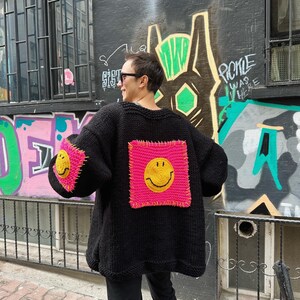 Puede incluir: Un c&aacute;rdigan negro de punto con parches cuadrados de color rosa ne&oacute;n con caras sonrientes amarillas. Las mangas tambi&eacute;n tienen los mismos parches de caras sonrientes. El c&aacute;rdigan se usa al aire libre, con una pared cubierta de grafitis en el fondo.