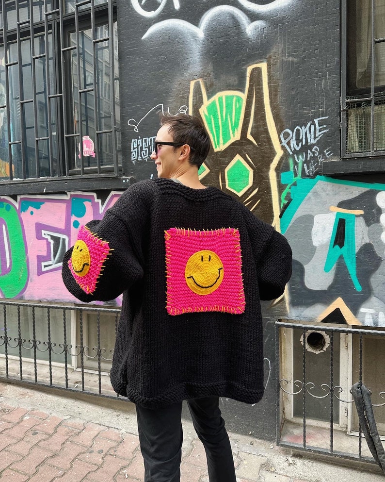 Puede incluir: Un c&aacute;rdigan negro de punto con parches cuadrados de color rosa ne&oacute;n con caras sonrientes amarillas. Las mangas tambi&eacute;n tienen los mismos parches de caras sonrientes. El c&aacute;rdigan se usa al aire libre, con una pared cubierta de grafitis en el fondo.