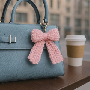 May include: A light blue handbag with a pink crocheted bow attached to the handle. The bow is made of a soft, textured yarn and hangs from a silver-colored clasp. A white paper coffee cup with a brown sleeve sits on a wooden surface.