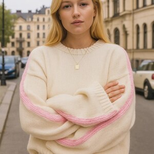May include: Cream-colored sweater with pink stripes on the sleeves. The sweater has a crew neck and long sleeves. A gold necklace with a square pendant is visible. The person is standing on a city street.