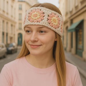 May include: A cream-colored crochet headband featuring three square floral designs with pink and yellow petals. The headband is worn by a person with long blonde hair and a pink shirt. The background shows a street scene with buildings.