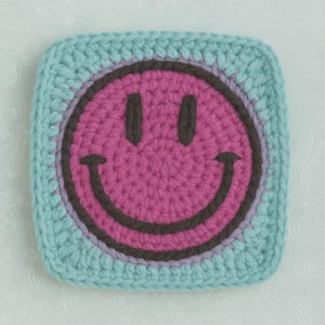 May include: A square, crocheted coaster with a smiley face design. The smiley face is pink with a dark brown outline and eyes, set against a light blue background. The coaster is handmade and has a textured appearance.