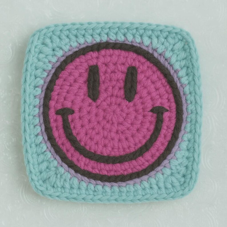 May include: A square, crocheted coaster with a smiley face design. The smiley face is pink with a dark brown outline and eyes, set against a light blue background. The coaster is handmade and has a textured appearance.