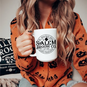 May include: A white ceramic mug with the text "Salem Broom Co. EST. 1692 Quality-Handcrafted" in black. The mug is held by a person wearing an orange sweater with black pumpkin patterns. The mug has a black witch graphic.