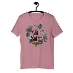May include: A heather pink t-shirt with a black and gray floral wreath design. The wreath includes a black moth and the text "Not every Witch lives in Salem".