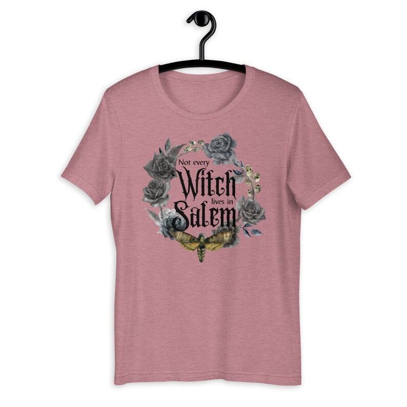May include: A heather pink t-shirt with a black and gray floral wreath design. The wreath includes a black moth and the text "Not every Witch lives in Salem".