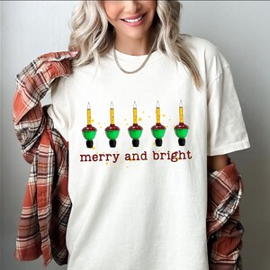 May include: White t-shirt with a graphic of five green and red Christmas lights with the text "merry and bright" underneath.
