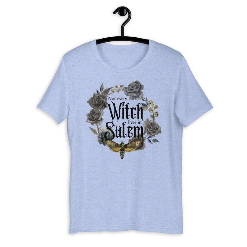 May include: Light blue t-shirt with a black and gray floral wreath design and a moth. The text "Not every Witch lives in Salem" is printed in black.
