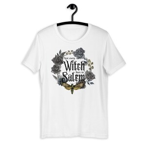 May include: White t-shirt with a black and gray floral wreath design. The wreath includes roses and a moth. The text "Not every Witch lives in Salem" is printed in black inside the wreath.