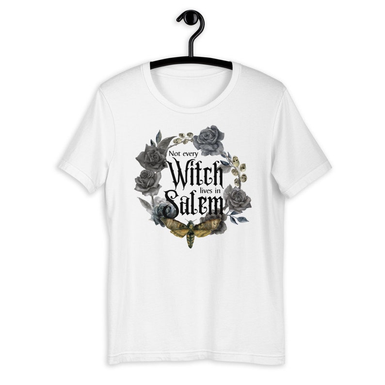 May include: White t-shirt with a black and gray floral wreath design. The wreath includes roses and a moth. The text "Not every Witch lives in Salem" is printed in black inside the wreath.