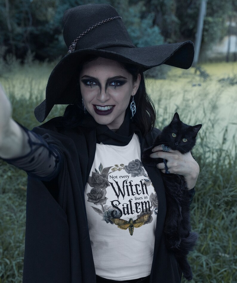 May include: A woman wearing a black witch hat and a white t-shirt with the text "Not every Witch lives in Salem" and a black cat in her arms. The t-shirt has a floral design with a moth.