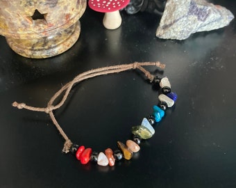 May include: A brown cord bracelet featuring an assortment of colourful gemstone beads, including red, orange, yellow, green, blue, and purple.