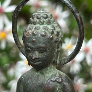 Peut inclure: Une figurine en métal patiné de couleur bronze représentant un Bouddha, dotée d'une texture vieillie et d'une auréole circulaire derrière la tête. La statue présente une coiffure traditionnelle ushnisha avec des petits reliefs arrondis.