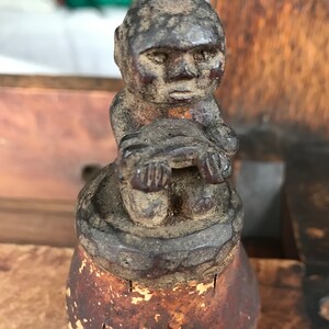 May include: A small, dark brown wooden figurine of a person sitting with their hands clasped in front of them. The figurine is carved with simple features and is mounted on a wooden base.