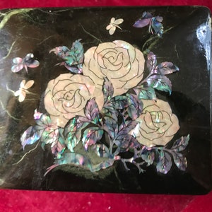 Pode incluir: Uma caixa lacada de preto com um design floral feito de madrepérola. O design apresenta três rosas brancas e folhas com tons azuis e verdes iridescentes. Há também quatro pequenas borboletas no design.