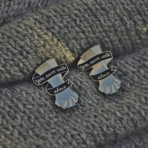May include: Two silver pins with black and white enamel. Each pin features a hand holding another hand with a banner that reads "You never walk alone".