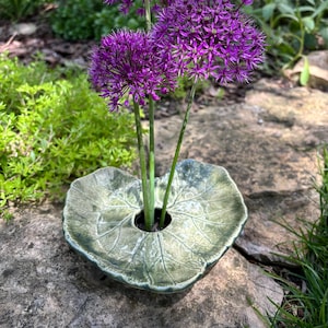 Puede incluir: Tres vibrantes flores de allium moradas con cabezas esféricas y tallos verdes delgados están dispuestas en un jarrón de cerámica con forma de hoja verde grande y texturizada. El jarrón presenta venas detalladas y una abertura central.