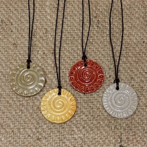 May include: Four ceramic pendant necklaces in shades of beige, yellow, red, and white. Each pendant is round with a spiral design and a brown leather cord. The necklaces are displayed on a textured, neutral-colored surface.