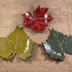 Puede incluir: Tres platos de cerámica con forma de hoja en rojo, verde y gris. Los platos están hechos para parecer hojas reales con venas y un tallo.