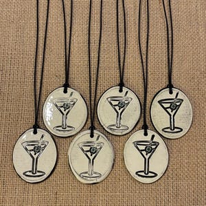 Puede incluir: Seis colgantes redondos de cerámica blanquecina con un diseño de copa de martini negra. Cada colgante cuelga de un cordón negro. Las copas de martini tienen una aceituna verde y una pajita.