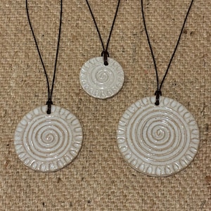 Puede incluir: Tres collares con colgantes de cerámica blanquecina y cordones de cuero marrón. Cada colgante presenta un diseño en espiral en el centro y un borde punteado. Los colgantes varían en tamaño.