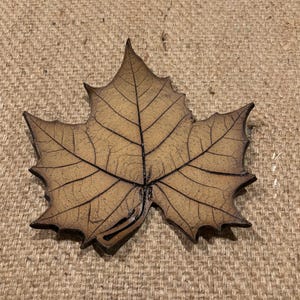 Pode incluir: Um prato decorativo em forma de folha com um padrão de veias detalhado. O prato é de cor castanha quente com detalhes em castanho escuro, assemelhando-se a uma folha caída. O prato em forma de folha é provavelmente feito de cerâmica ou metal.