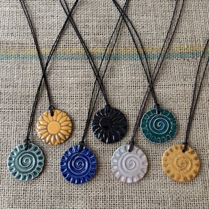 Op de afbeelding: Een verzameling keramische hangerkettingen met een cirkelvormig ontwerp. De hangers zijn in verschillende kleuren, waaronder geel, zwart, blauw en wit, en zijn geregen aan zwarte koorden. De kettingen worden gepresenteerd op een gestructureerd, neutraal gekleurd oppervlak.