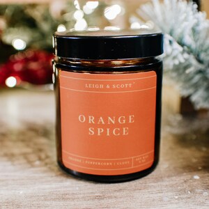 Puede incluir: Un tarro de vela de vidrio marr&oacute;n con tapa negra. La etiqueta es naranja y dice "Leigh & Scott" y "Orange Spice".