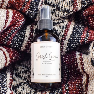Può includere: Una bottiglia di vetro marrone con un'etichetta bianca che recita "Leigh & Scott Fresh Linen Deodorizing Room Spray Made with Essential Oils 4 oz". La bottiglia è appoggiata su una coperta a strisce rosse e bianche.