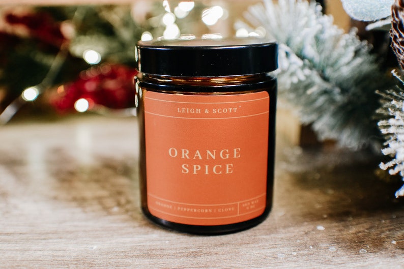 Puede incluir: Un tarro de vela de vidrio marr&oacute;n con tapa negra. La etiqueta es naranja y dice "Leigh & Scott" y "Orange Spice".