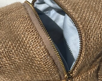 May include: A tan burlap pouch featuring a coarse, woven texture and a gold-toned metal zipper closure. The interior of the pouch is lined with a smooth, light blue fabric, visible through the partially opened zipper opening at the top.