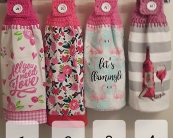May include: Four hanging kitchen towels with crocheted pink tops and white buttons. Towel 1 has pink hearts and text "All you need is love". Towel 2 has floral patterns. Towel 3 has flamingos and text "let's flamingle". Towel 4 has wine and stripes.